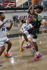 A basketball player in a black and green uniform jumps to shoot the ball as two defenders in white uniforms attempt to block him. The action takes place on an indoor court with spectators seated in the background.