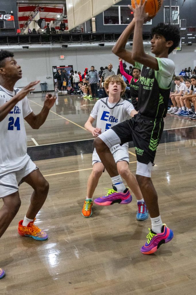 A basketball player in a black and green uniform jumps to shoot the ball as two defenders in white uniforms attempt to block him. The action takes place on an indoor court with spectators seated in the background.
