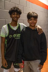 Two young people stand side by side in a hallway with gray brick walls. One is wearing a black and green sports jersey and shorts, while the other is in a black hoodie and gray pants. They both have short, dark hair.
