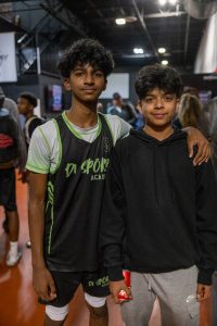 Two boys pose indoors at a sports facility. One wears a black and green sports jersey with "DA SPORTS ACADEMY" printed on it. The other wears a black hoodie and gray pants. One has an arm around the other's shoulder.