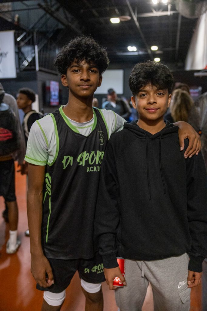Two boys pose indoors at a sports facility. One wears a black and green sports jersey with "DA SPORTS ACADEMY" printed on it. The other wears a black hoodie and gray pants. One has an arm around the other's shoulder.