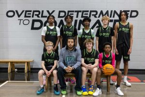 A basketball team of eight players wearing black uniforms pose with their coach, who is wearing a gray D1 Sports Academy hoodie. One player sits holding an orange basketball. The wall behind them features the words "Overly Determined to Dominate.