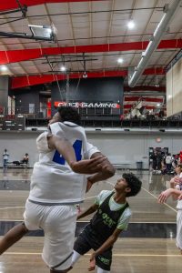 A basketball game in an indoor court. A player in a white jersey jumps to block or shoot, while a player in a black and green jersey reaches up. The court has spectators and a red steel beam ceiling with "Gym Gaming" written on a wall.