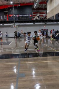 Two basketball players on a court. The player in black drives towards the basket, guarded by a player in white. Spectators watch from the sidelines. A basketball hoop and "ONExGAMING" banner are visible in the background.