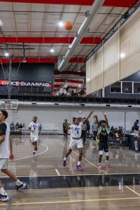 A basketball game in an indoor court with players in white jerseys defending against a player in a black jersey. The ball is in mid-air toward the basket, and a referee stands nearby. Spectators can be seen in the background.