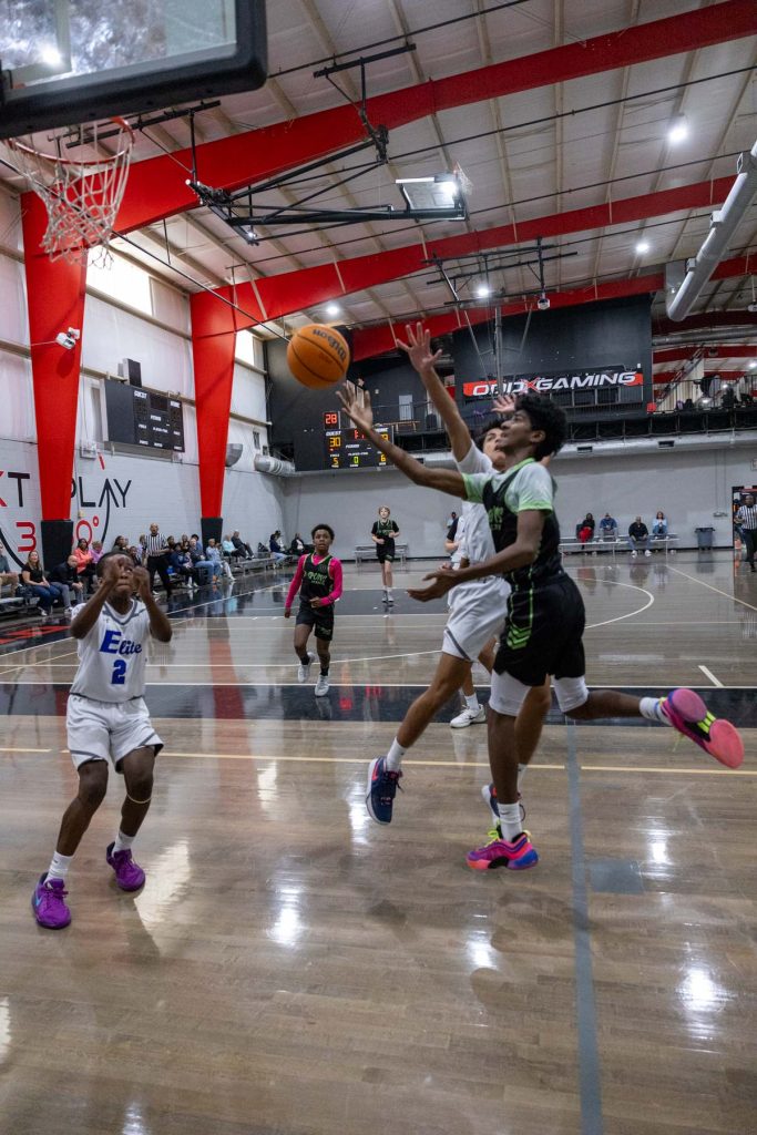 Two basketball players leap towards the hoop, each trying to score or block the shot. They are on an indoor court with spectators sitting in the background. A digital scoreboard is visible above them.